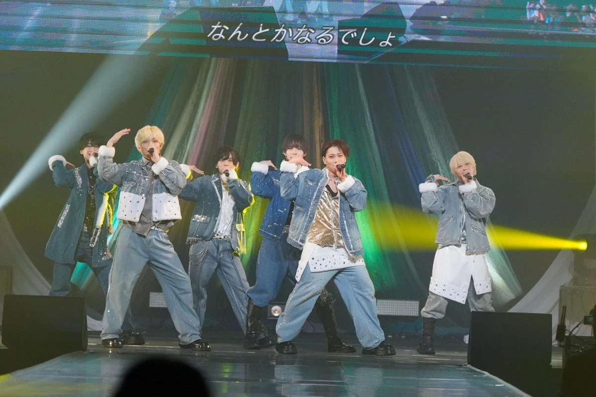 ステージ上で6人の男性アイドルグループが歌唱パフォーマンスをしている。デニムとファーを組み合わせたきらびやかな衣装をまとい、マイクを手に歌っている。背景のスクリーンには「なんとかなるでしょう」の文字が表示され、華やかな照明が演出を彩っている。