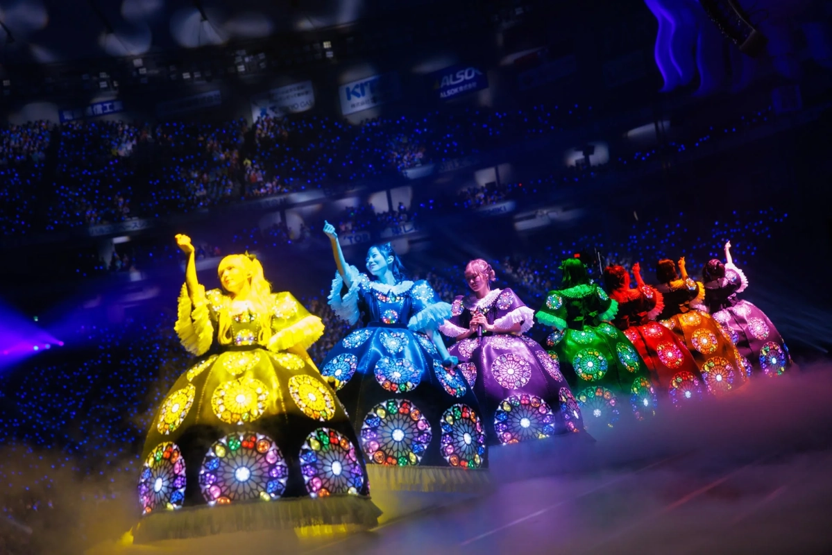 ステンドグラスのような光るカラフルなドレスをまとったパフォーマーたちが、青いペンライトが輝くアリーナで華麗なパフォーマンスを披露しています。幻想的な煙がステージを彩り、会場は熱気に包まれています。