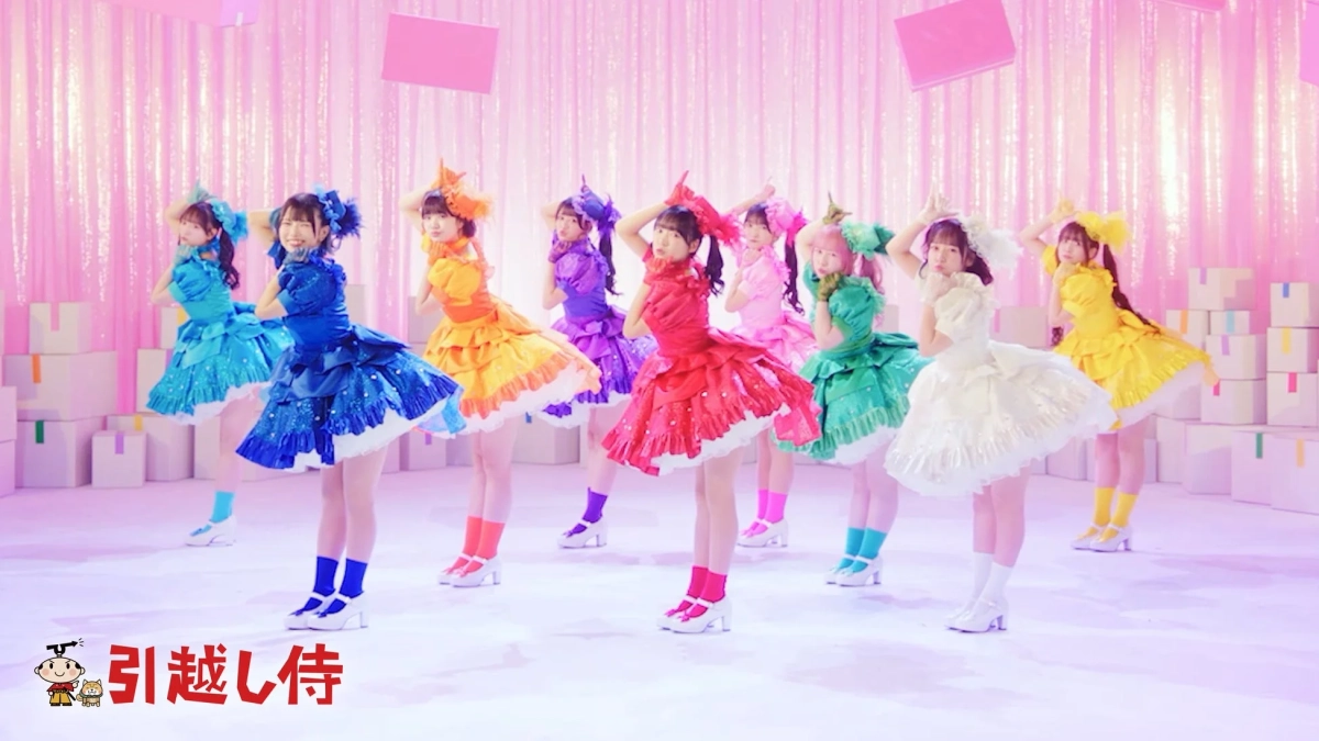 カラフルなドレスを身につけた複数の女性が、ピンクのきらびやかな背景と段ボールが積まれた空間でポーズをとっています。明るく華やかな雰囲気で、ダンスパフォーマンスの一場面のようです。左下には「引越し侍」のロゴが見えます。