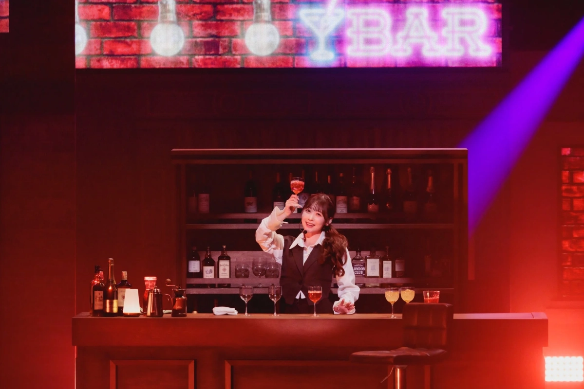 赤い照明が印象的なバーカウンターで、笑顔の女性バーテンダーがカクテルグラスを掲げている