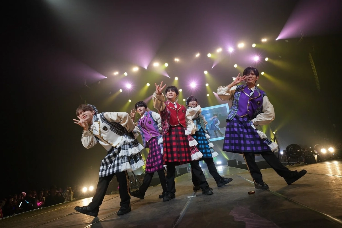 明るいステージライトの下で、5人の男性アイドルグループがパフォーマンスをしている様子。カラフルなチェック柄の衣装を身につけ、皆がピースサインをして笑顔を見せている。ライブコンサートの活気ある雰囲気が伝わる一枚。