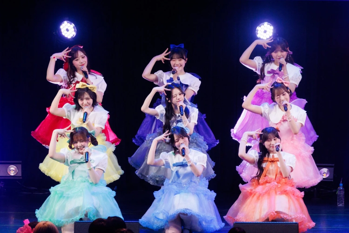 カラフルなドレスをまとった9人の女性アイドルグループが、ステージ上でマイクを持ち、様々なポーズを取りながらパフォーマンスをしている様子です。明るい笑顔で観客を魅了しているライブの一幕と見られます。