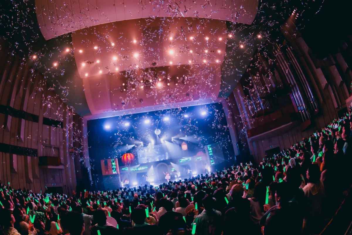 ライブ会場で多くの観客がペンライトを振る中、ステージではパフォーマーが歌い、上からは紙吹雪が舞い散る賑やかなコンサートの様子