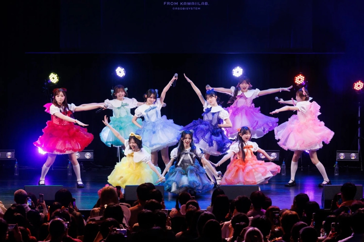カラフルなフリルドレスをまとった女性アイドルグループがステージで歌い踊るライブパフォーマンスの様子です。多数の観客が見守り、会場は熱気に包まれています。