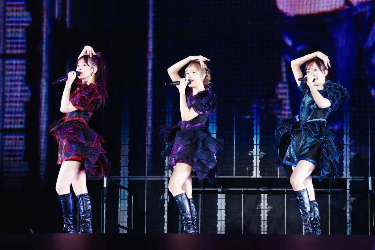 華やかな衣装を身につけた3人の女性アーティストが、マイクを手にステージでパフォーマンスしている様子。青と紫の照明が印象的なライブコンサートの瞬間を捉えた写真です。