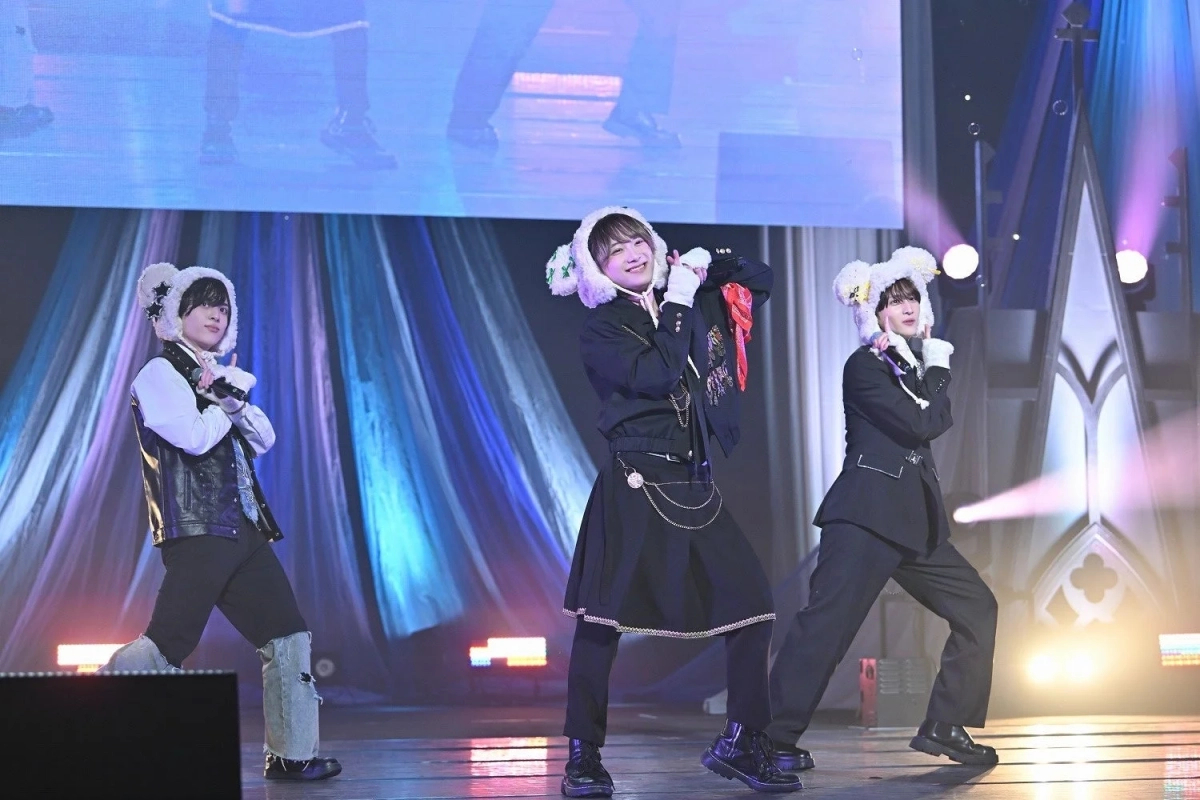 カラフルな照明が当たるステージ上で、白いアニマルハットと個性的な衣装を身につけた3人の男性パフォーマーが、楽しそうにポーズを取っている様子。ライブやコンサートの一場面を捉えた写真です。