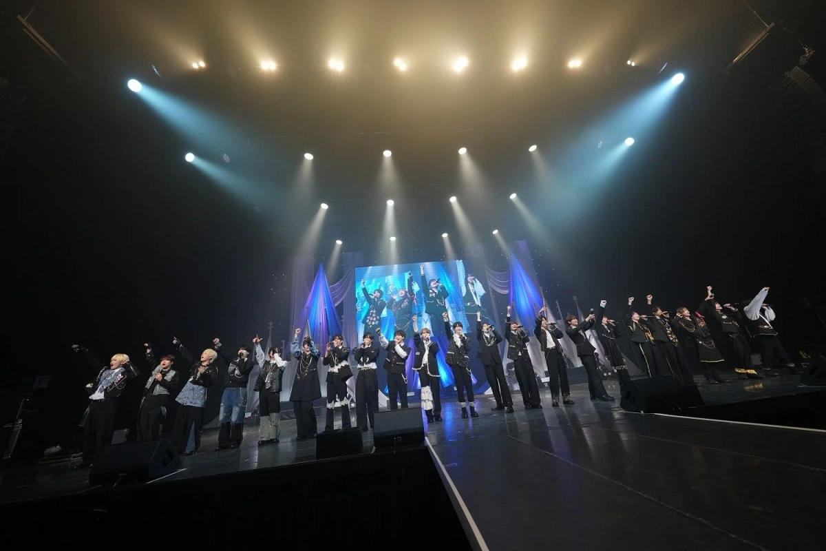 多数の男性パフォーマーがステージ上で歌い、観客に向かって拳を突き上げているライブコンサートの様子。明るいスポットライトと背景の大型スクリーンが印象的で、熱気あふれるパフォーマンスが繰り広げられています。
