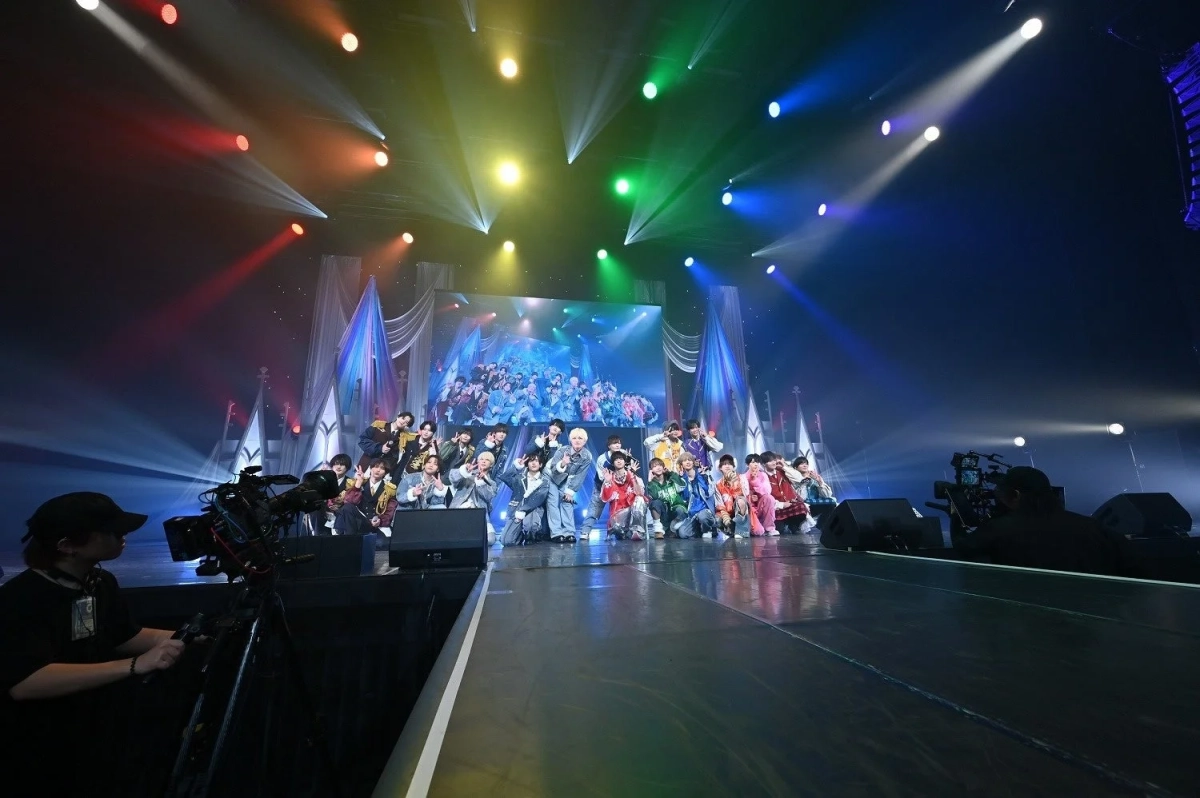 カラフルな衣装を身につけた大勢のパフォーマーが、華やかな照明が輝くステージ上でポーズをとっています。背景には大型スクリーンがあり、手前には撮影中のカメラとスタッフが写り込んでいます。