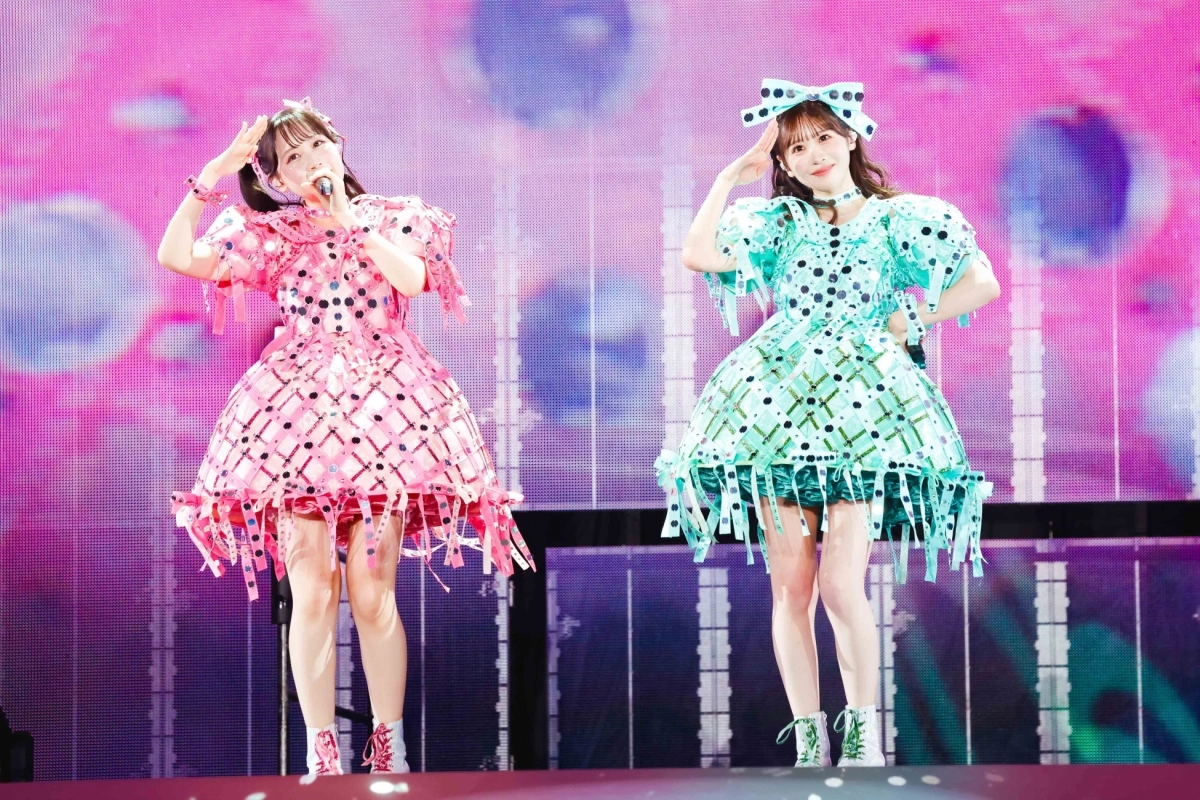 華やかなステージでパフォーマンスを行う2人の女性アイドル。ピンクと水色の特徴的な衣装を身につけ、リボンや飾りが目を引きます。笑顔でポーズをとり、ライブの楽しさが伝わる一枚です。