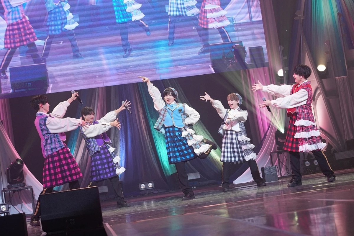 カラフルなチェック柄の衣装を身につけた5人の男性アイドルが、明るいステージでマイクを手に歌い踊っている様子。活気あふれるライブパフォーマンスの一場面です。