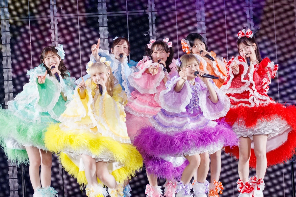カラフルでフリルの多い衣装を身につけた7人の女性アイドルグループが、ステージ上で笑顔でパフォーマンスしている様子。それぞれマイクを持ち、楽しそうに歌っており、華やかなコンサートの雰囲気が伝わります。