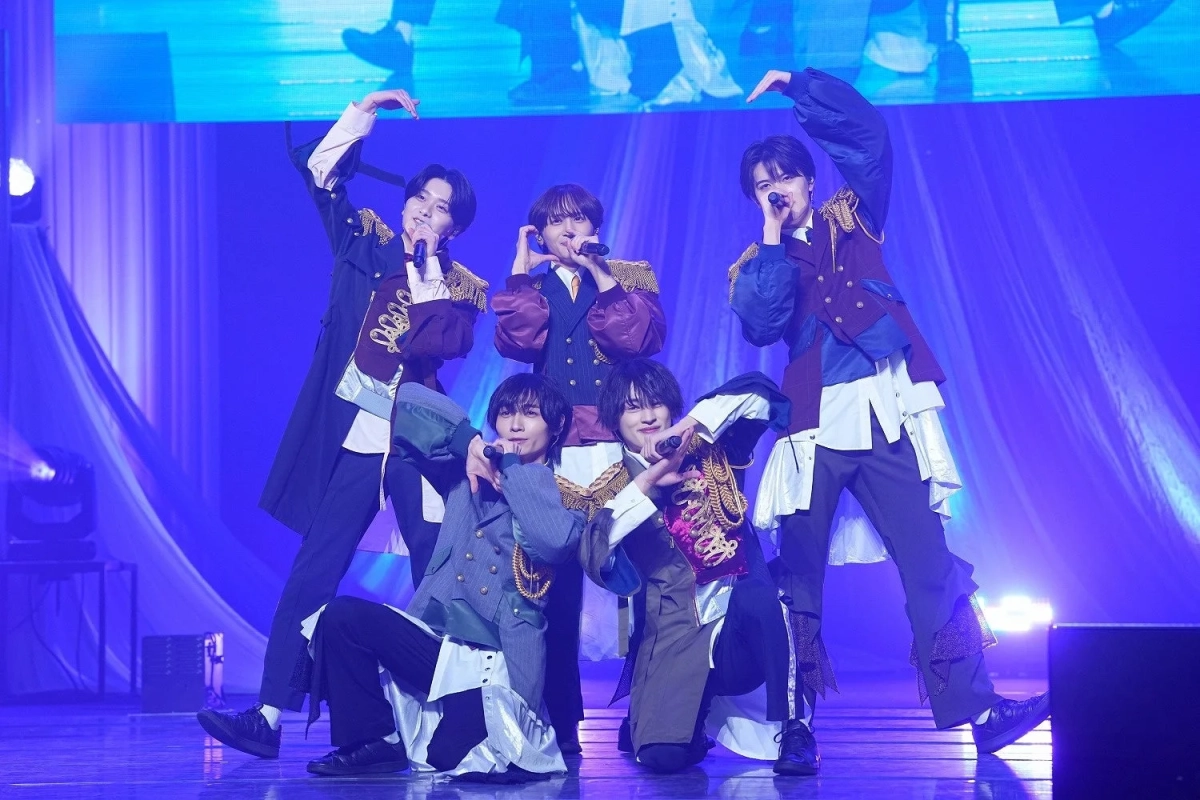 青い照明が印象的なステージで、華やかな衣装をまとった5人の男性アイドルグループがマイクを手にパフォーマンスしています。ポーズを決めており、ライブの熱気が伝わる一枚です。