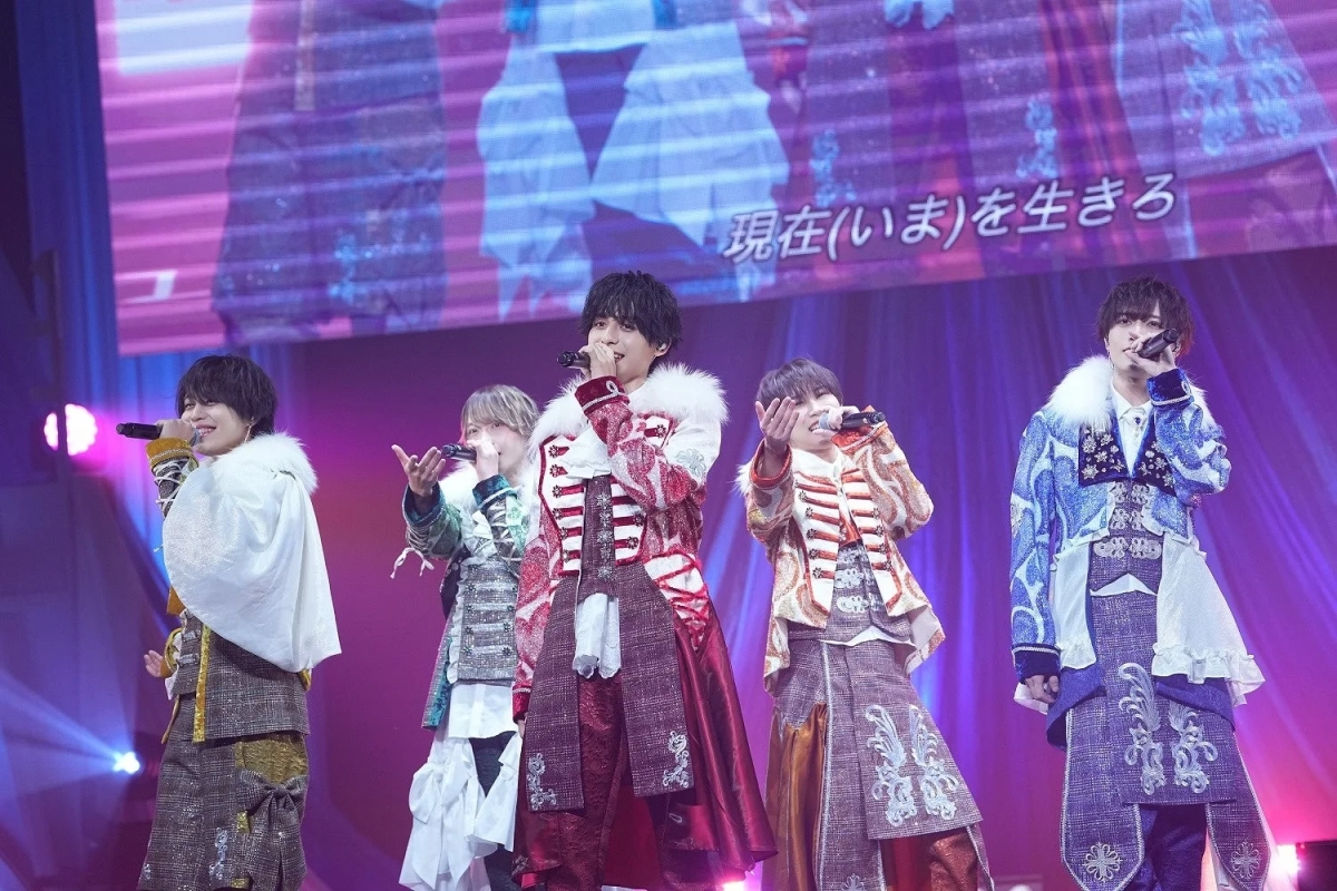 豪華な装飾の衣装をまとった5人の男性アイドルグループが、ステージ上でマイクを手に熱唱しているライブパフォーマンスの様子。背景のスクリーンには「現在(いま)を生きる」というメッセージが映し出されている。