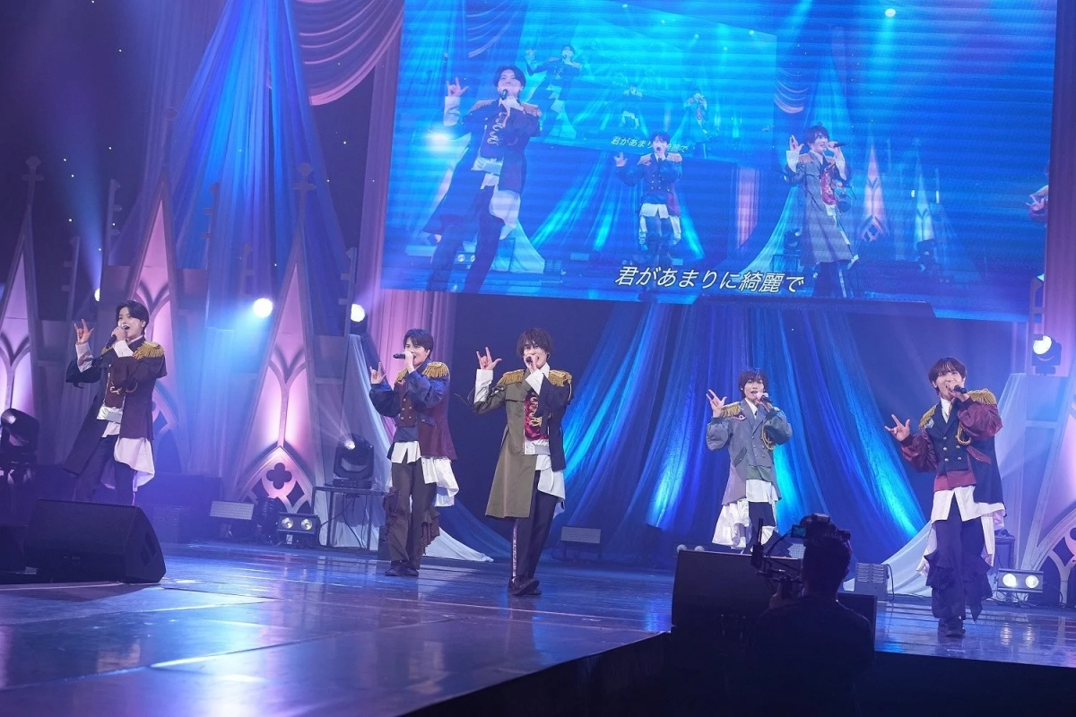 豪華な衣装をまとった5人の男性パフォーマーが、青と紫の照明が照らすステージで歌唱パフォーマンスを行っています。背景の大型スクリーンには歌詞とメンバーの姿が映し出されており、熱気あふれるライブの様子を捉えています。