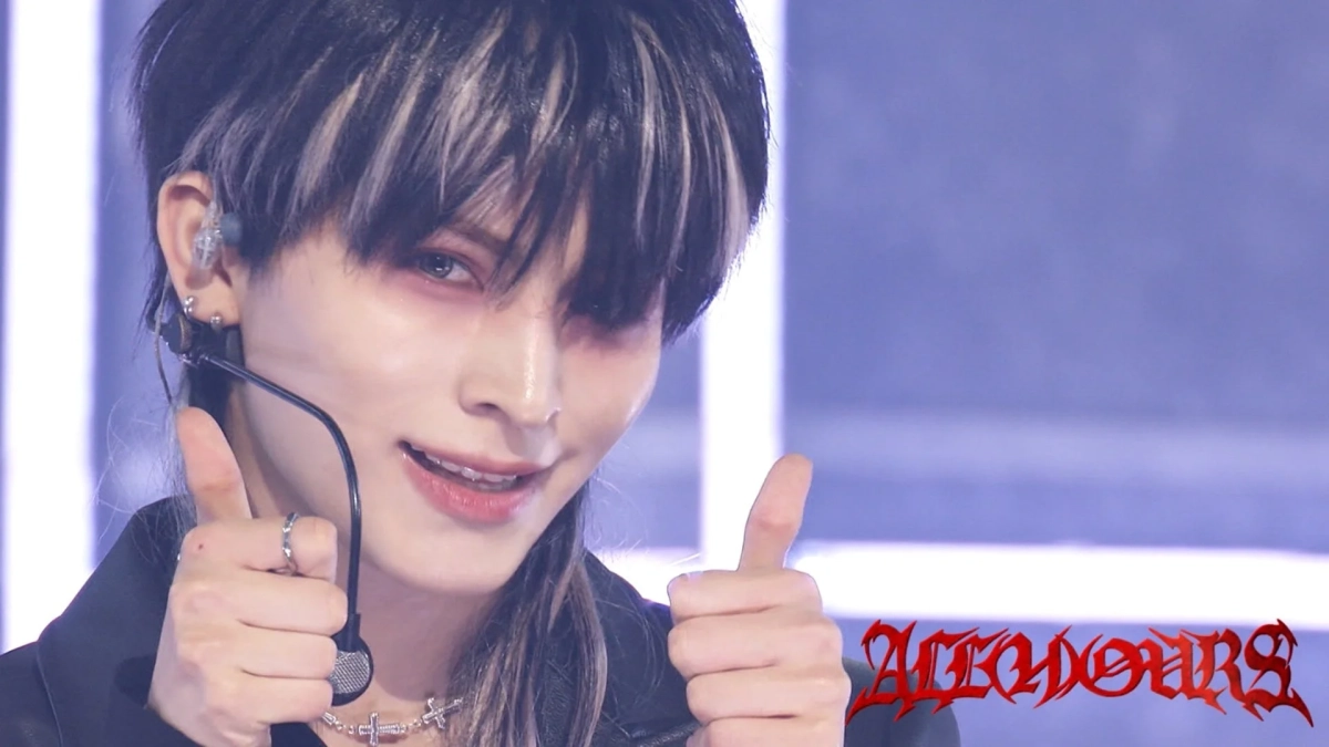 独特なメイクとツートンヘアの男性が、ステージ上で笑顔でダブルサムズアップをしている様子。