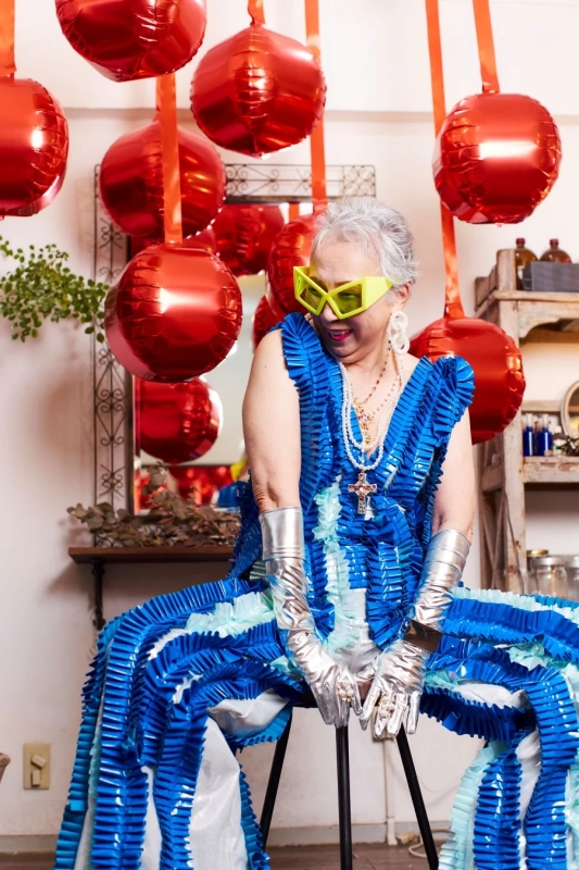 鮮やかな青いフリルとメタリックな手袋、ネオンイエローのサングラスを身につけた高齢の女性のポートレート