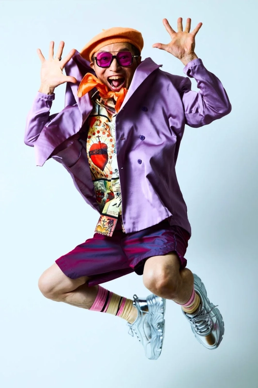 カラフルで個性的なファッションに身を包んだ男性が、満面の笑みでジャンプしている瞬間