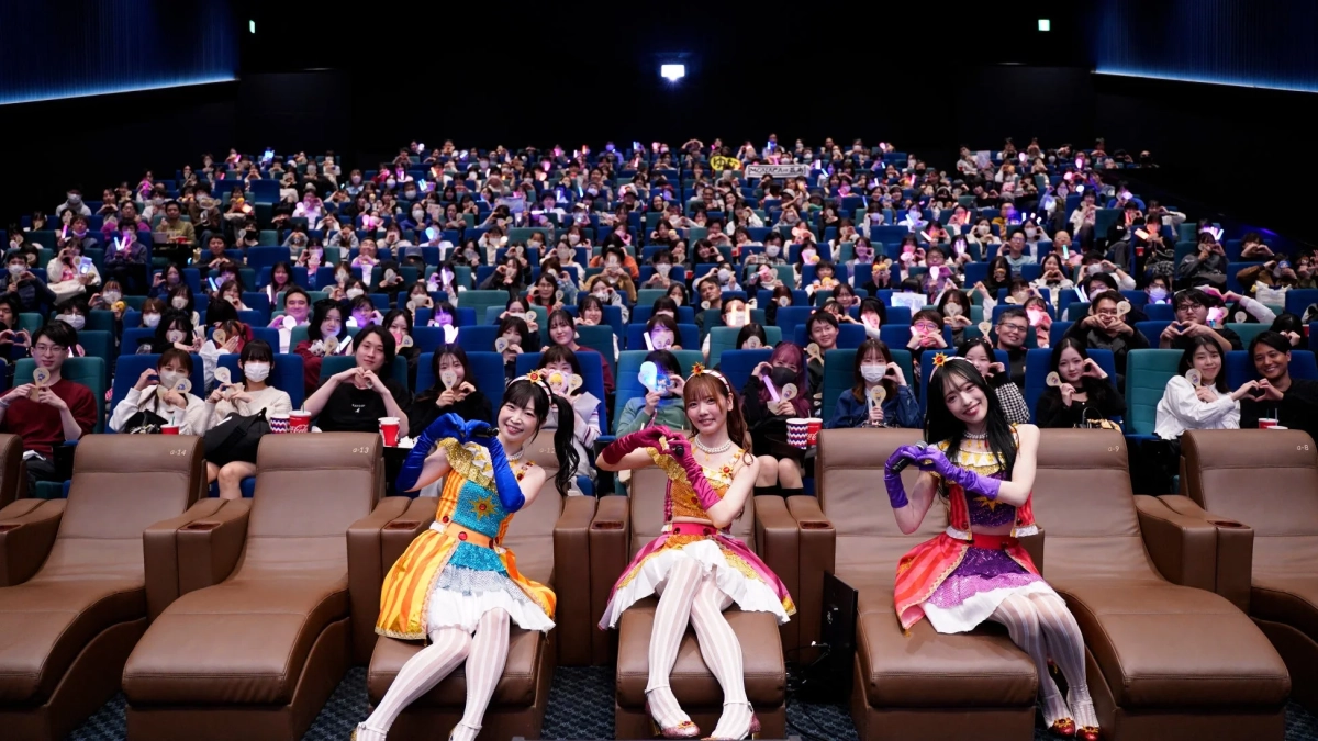 カラフルな衣装を着た3人のアイドルが最前列に座り、映画館のような会場で多くのファンがハートマークやペンライトで応援しているイベントの様子。