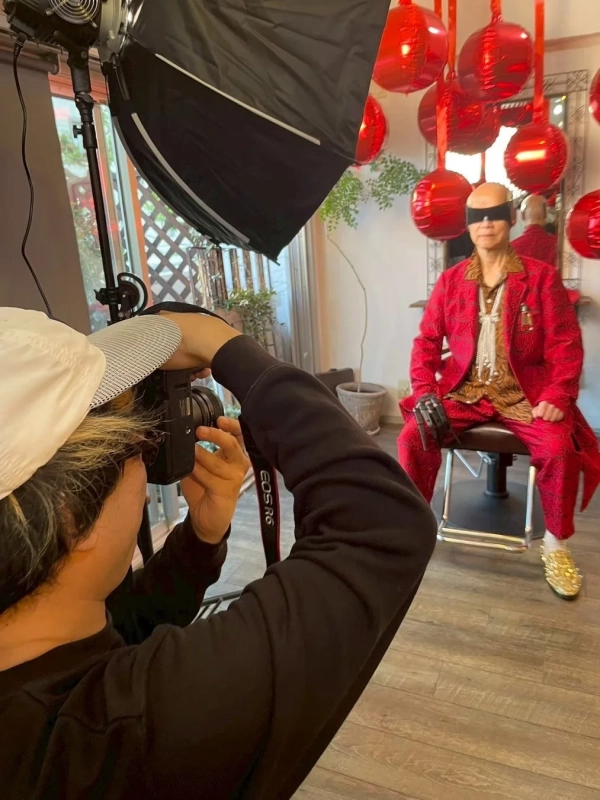 赤いスーツにブラインドと金色のスパイクシューズを履いた男性が椅子に座り、カメラマンが撮影している様子