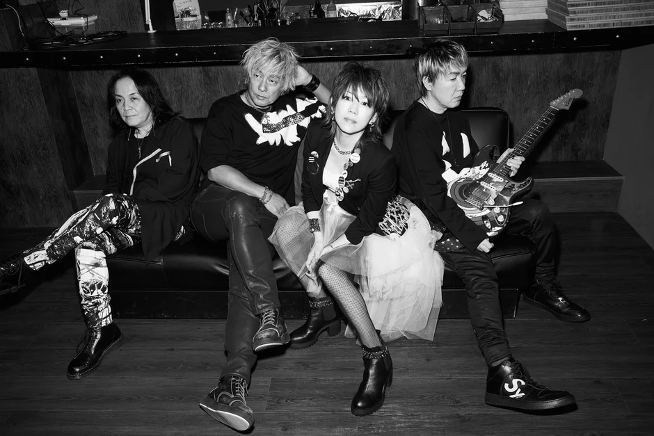 ソファに座ってポーズをとる4人組の男女混合バンドの白黒写真。一人がエレキギターを抱えており、ロックな雰囲気が漂っている。