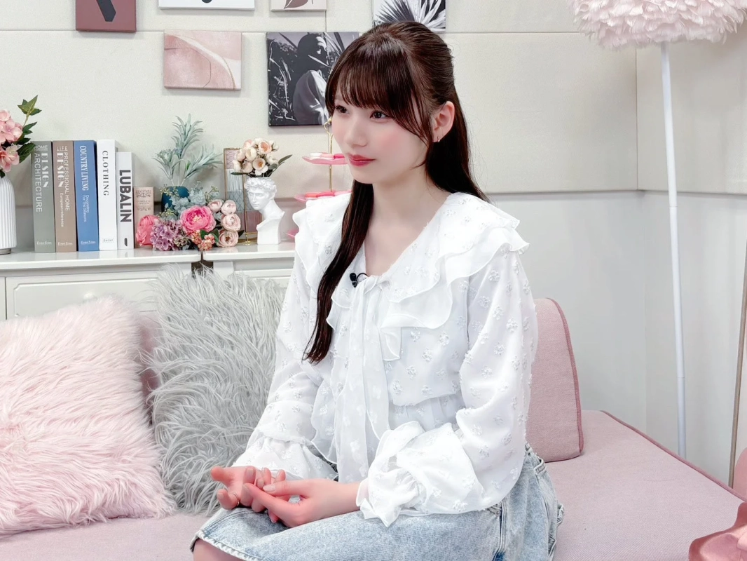 白いフリルブラウスとジーンズ姿の若い女性が、ピンクのソファに座って横を向いている