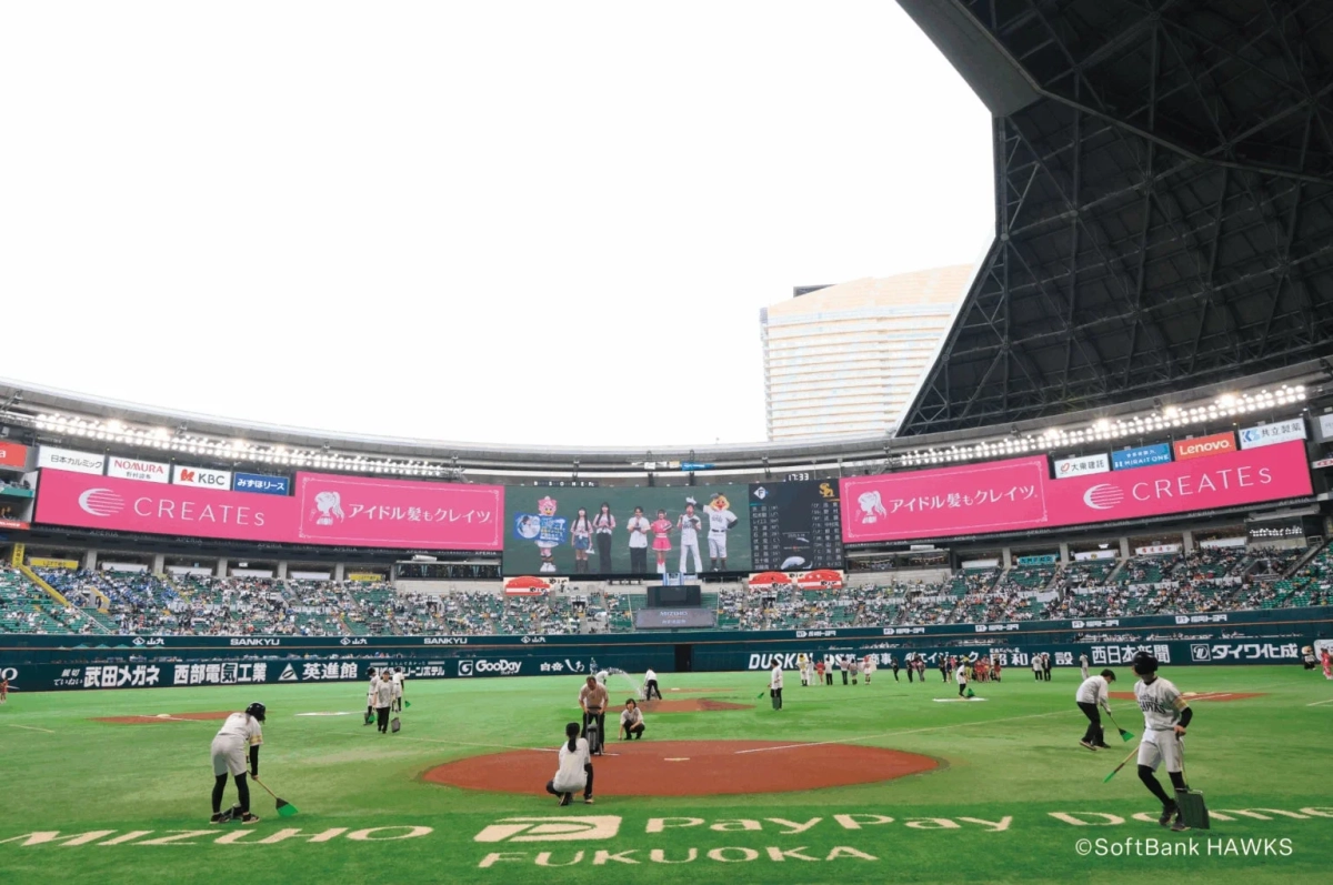野球場でグラウンド整備が行われている様子。大型ビジョンには広告やイベントが映し出され、観客席には多くのファンが見守っている。PayPayドームと福岡ソフトバンクホークスのロゴが見える。