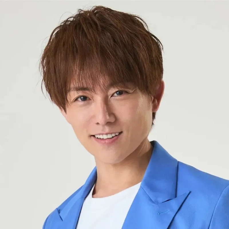 白い背景の前で、青いジャケットと白いTシャツを着用した茶髪の男性が笑顔でカメラを見ているポートレート写真です。