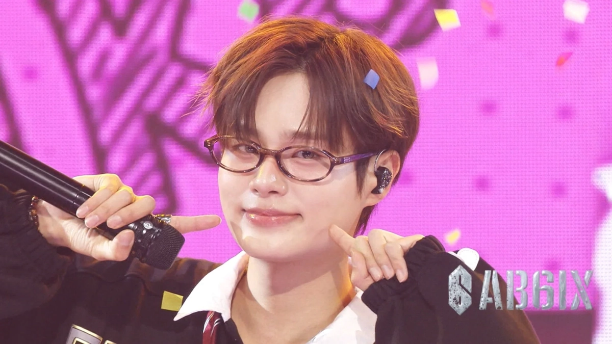 ピンク色のステージでパフォーマンス中の男性アイドルが、眼鏡をかけ、マイクを持ちながら頬を指して笑顔を見せています。