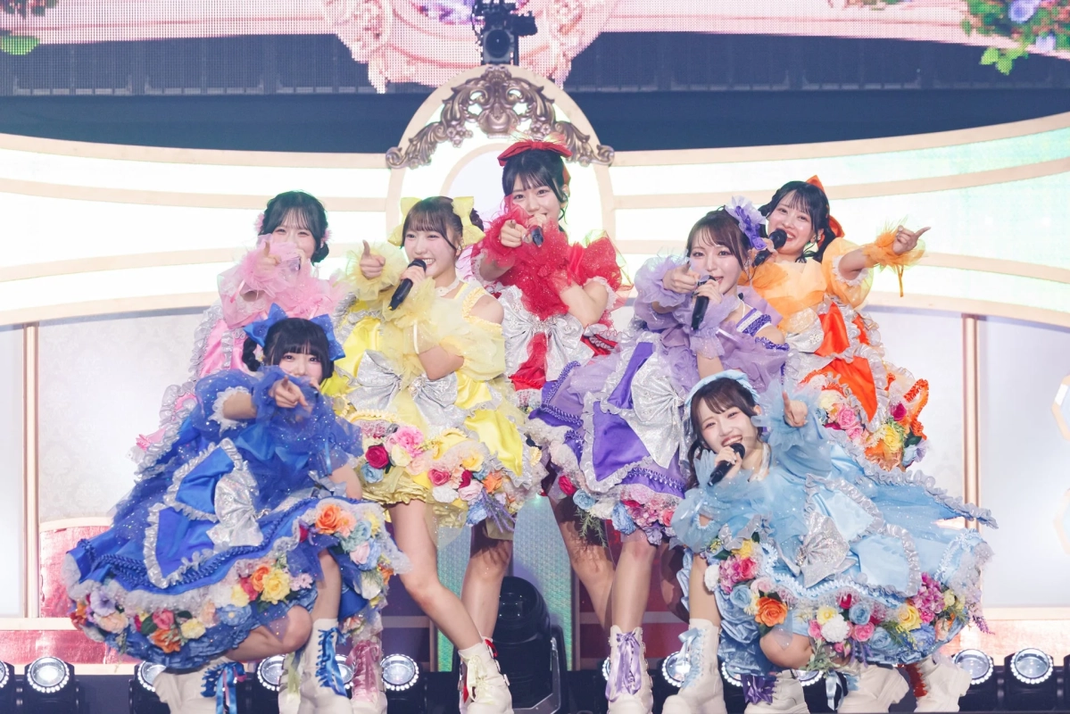 華やかな衣装を身につけた7人の女性アイドルグループが、ステージ上で笑顔でパフォーマンスしている様子。カラフルなドレスにはたくさんの花が飾られ、ライブの楽しそうな雰囲気が伝わってきます。