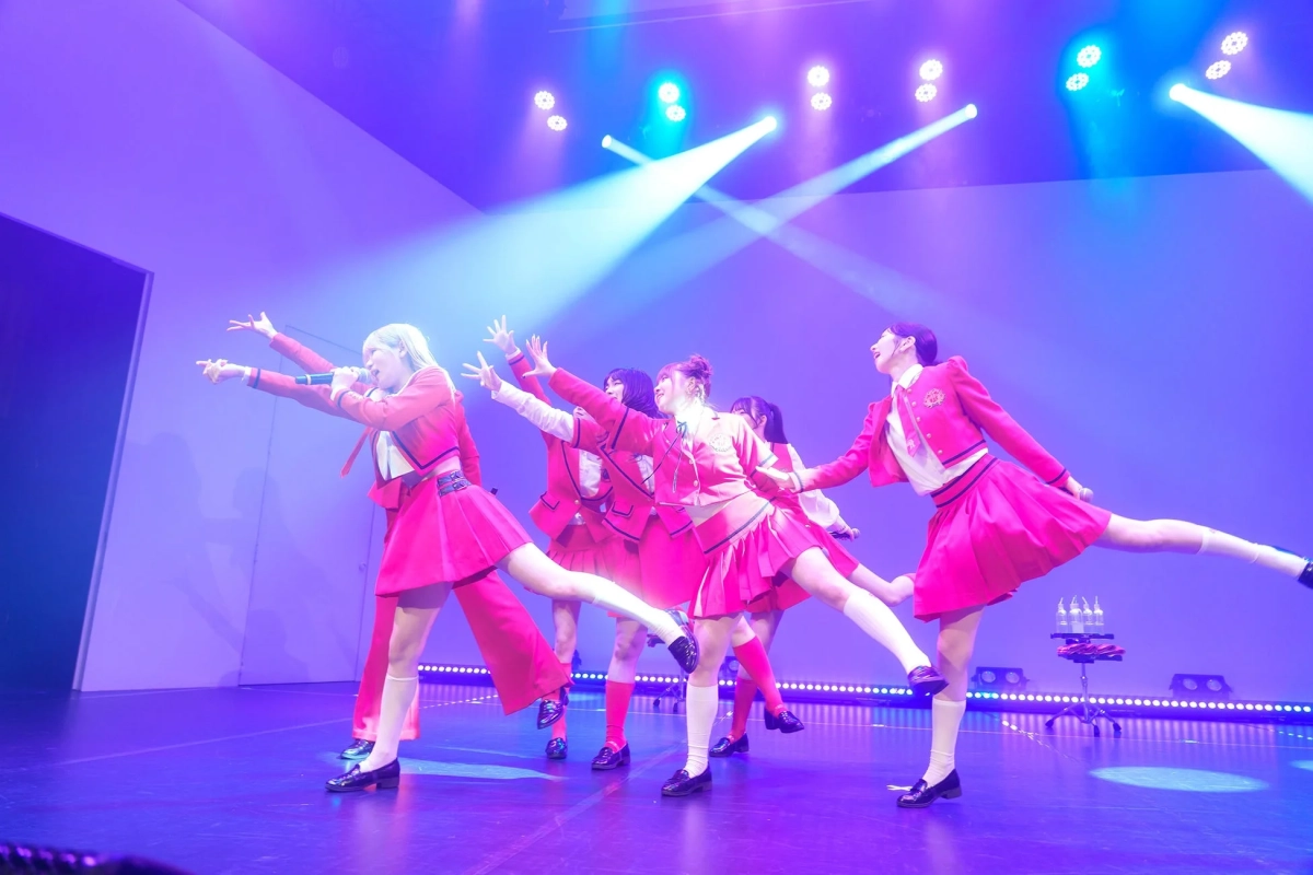 鮮やかなピンクの制服を着た女性アイドルグループが、ステージ上でスポットライトを浴びながらダイナミックなダンスパフォーマンスを披露しています。
