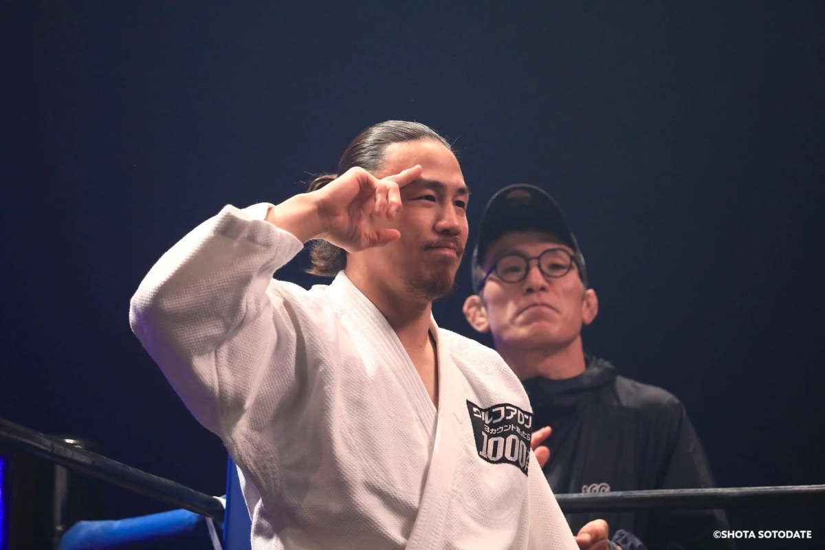 白い柔術着を着た男性がリング上でポーズをとっており、その後ろには黒い服を着た男性が立っている。格闘技イベントの一場面で、スポンサーロゴも見える。