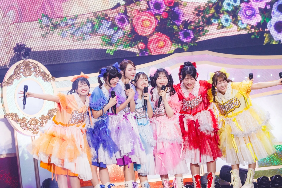 カラフルなフリル衣装を着た7人組の女性アイドルグループが、ステージ上で笑顔でパフォーマンスしているライブ風景。背景には花柄の装飾が施されている。