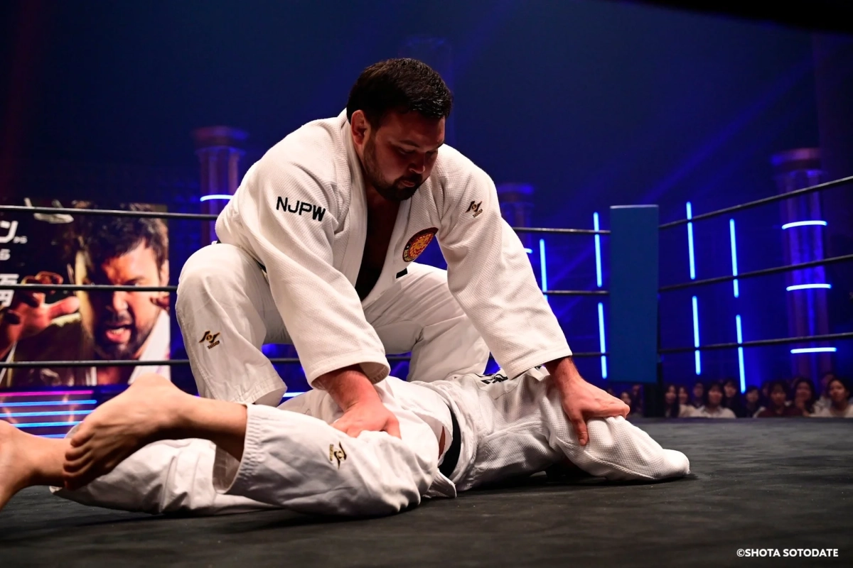 白い道着を着た二人の男性がリング上で寝技を繰り広げている格闘技の試合風景です。一人の選手がもう一人の選手を抑え込んでいます。