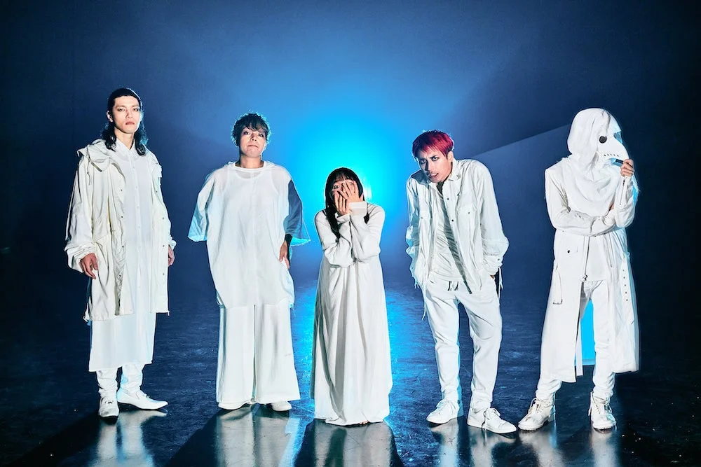 青い光が当たる暗い背景の中、白を基調とした個性的な衣装を身につけた5人が一列に並んで立っています。顔を覆う人物や仮面をつけた人物もおり、神秘的で芸術的な雰囲気を醸し出しています。