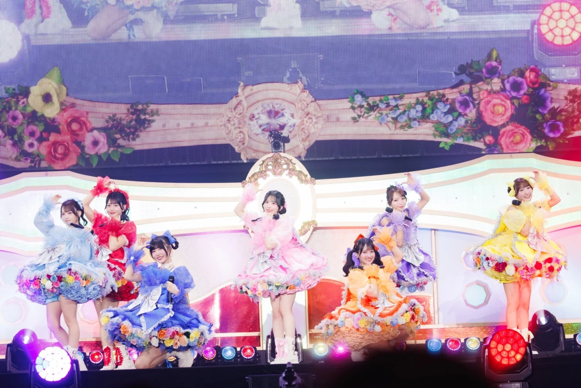 華やかなステージで、カラフルなフリルと花飾りの衣装をまとった複数の女性アイドルがパフォーマンスしている様子。背景には大きなスクリーンに花の模様が映し出され、スポットライトが煌めくライブ会場の熱気が伝わる一枚。