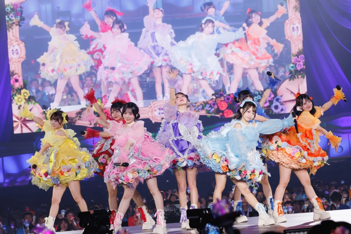 カラフルな花柄の衣装をまとった女性アイドルグループが、明るいステージでパフォーマンスを披露している様子。観客が見守る中、メンバーは笑顔で手を広げ、華やかなライブの雰囲気が伝わってきます。