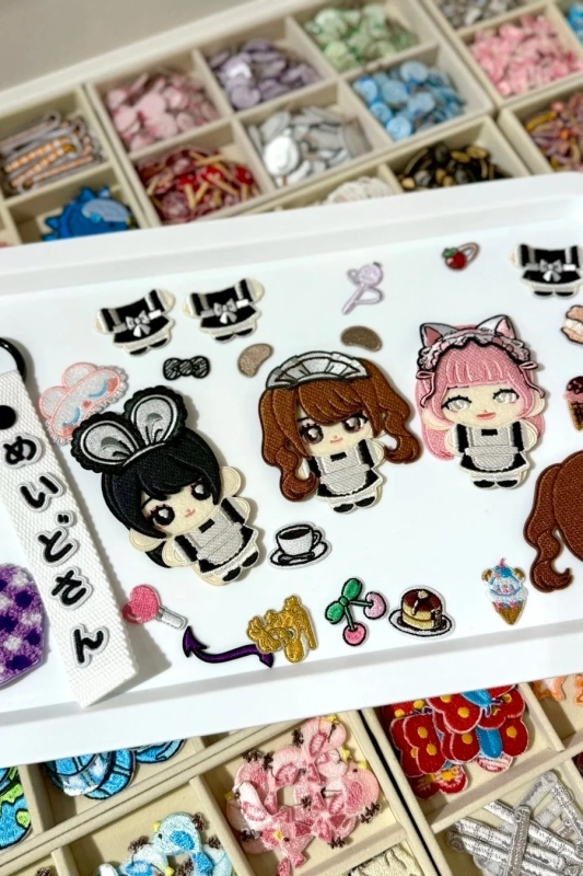 メイドキャラクターや可愛らしい食べ物などの刺繍ワッペンが白いトレイに並べられ、背景には整理された収納ボックスが見える