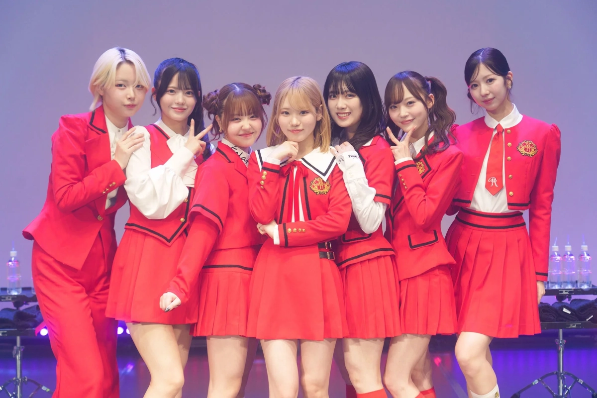 赤い制服を着た7人の女性アイドルグループがステージ上で笑顔でポーズをとっている集合写真です。