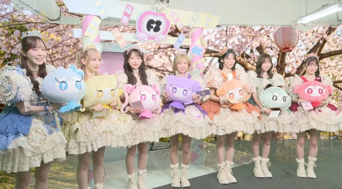 フリルとレースの豪華な衣装を着た若い女性たちが、それぞれ異なる色の可愛いキャラクターのぬいぐるみを抱えてステージに立っている。背景には桜の木と「F3」のロゴ、「祝」「祭」と書かれた装飾があり、華やかなイベントの様子が伺える。