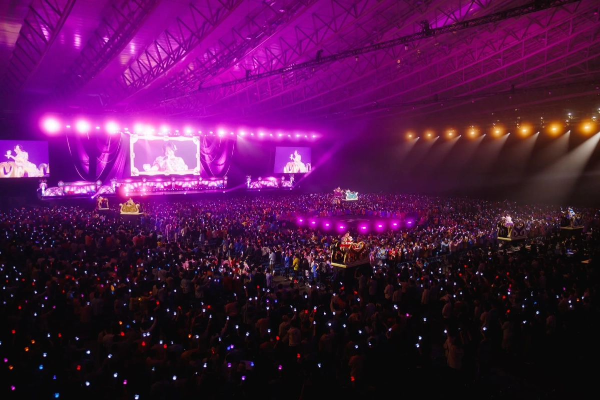 紫色の照明に包まれた広大なアリーナで、大勢の観客がペンライトを振って盛り上がるライブイベントの様子。巨大スクリーンにはパフォーマーが映し出され、フロートに乗った演者も客席を移動している。