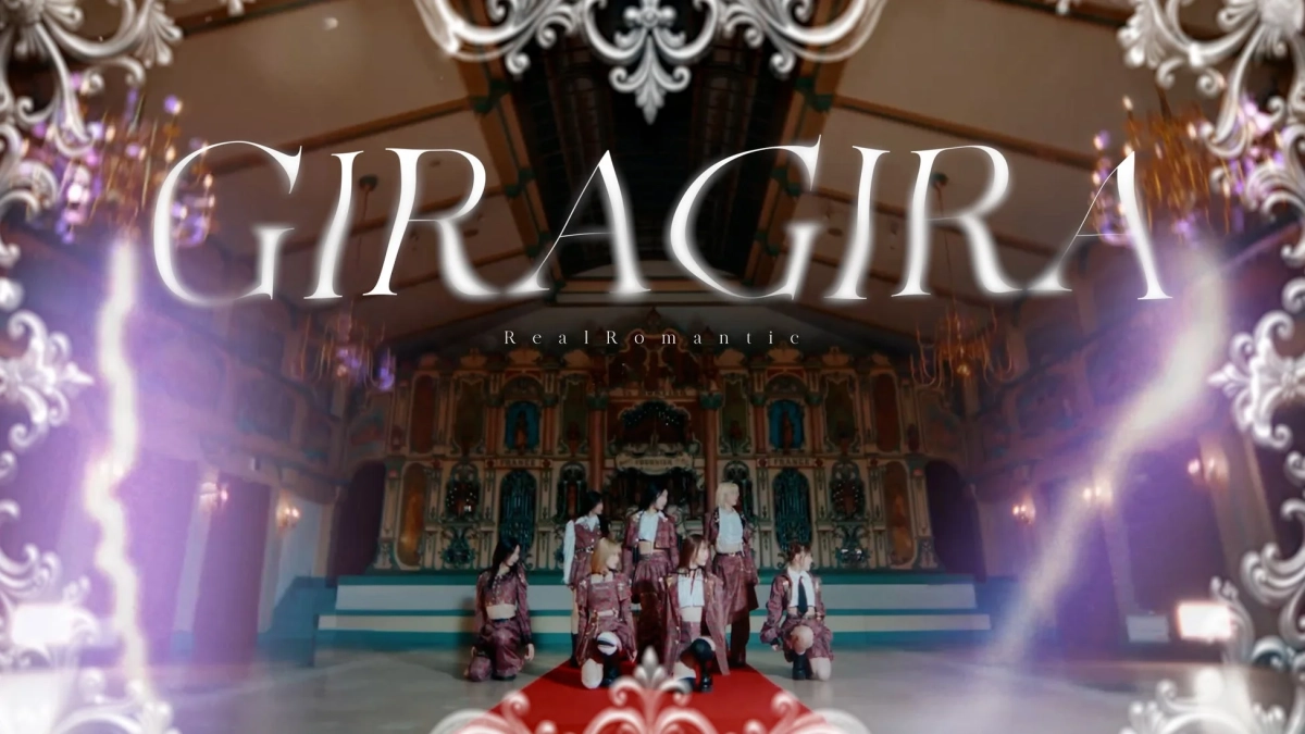 豪華な装飾が施されたステージで、チェック柄の衣装をまとった女性アイドルグループがポーズをとっている。中央には「GIRAGIRA」と「Real Romantic」の文字があり、ミュージックビデオの一場面のようだ。