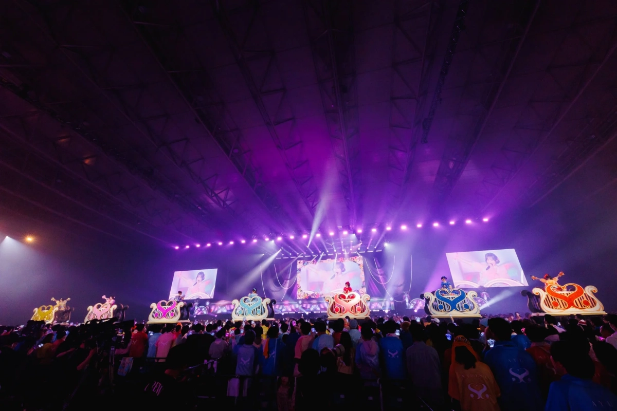 大規模なアリーナで行われているライブコンサートの様子。多数の観客がカラフルな照明と巨大スクリーン、そして豪華なフロートに乗ったパフォーマーのステージパフォーマンスを楽しんでいる。会場全体が紫色の光に包まれ、熱気に満ちている。