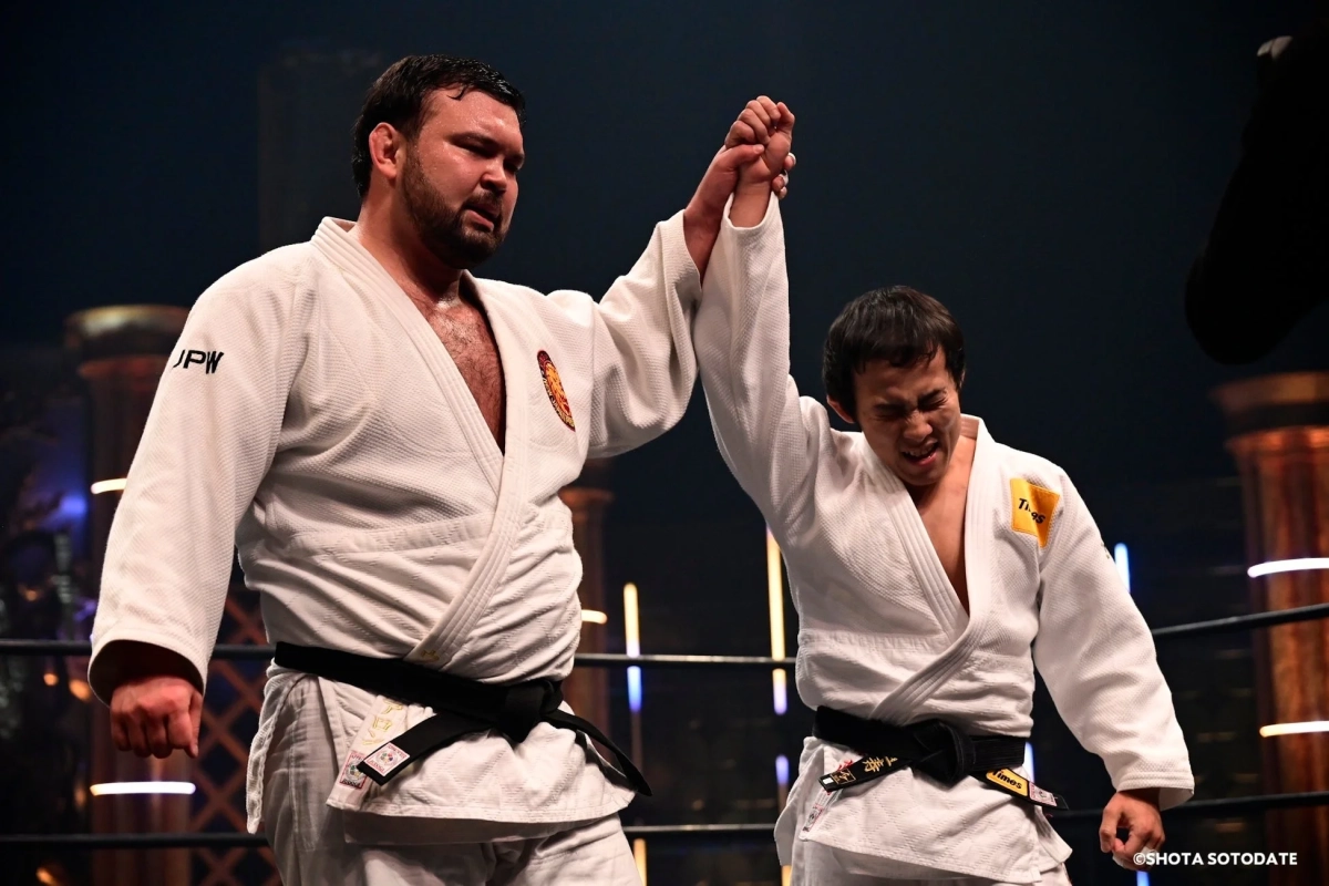 柔道着を着用した二人の男性がリング上で、片方がもう片方の腕を高く掲げている。試合後の勝利セレモニーの様子で、勝者と敗者の表情が対照的である。