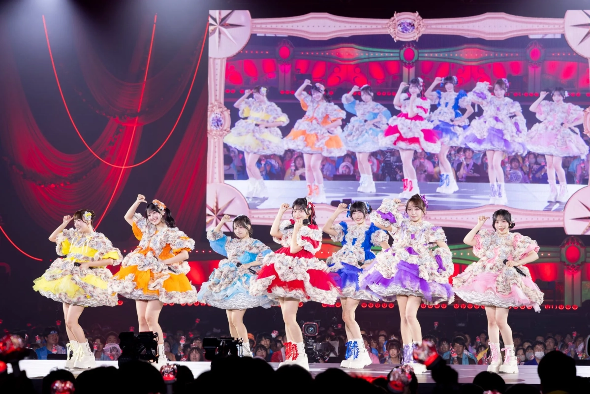 複数の女性アイドルグループがカラフルなフリルの衣装を着てステージでパフォーマンスしている様子。背景の大型スクリーンにも同じグループの映像が映し出され、観客も見える。ライブコンサートの熱気が伝わる一枚です。
