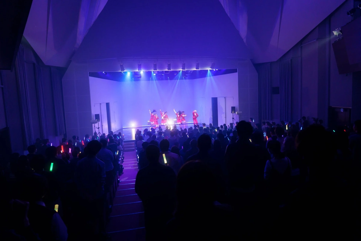 青い照明が当たるステージで複数のパフォーマーが演技しており、暗い客席では観客が色とりどりのペンライトを振っているライブ会場の様子。