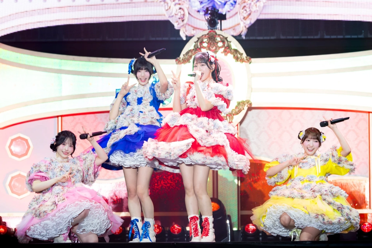 ステージ上で、カラフルなフリルと花の装飾が施されたドレスを着た4人の女性アイドルが笑顔でパフォーマンスしている様子。マイクを持ち、ピースサインをするなど、明るく楽しいライブの雰囲気です。