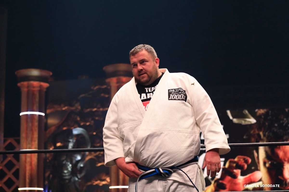 白い道着と青帯を着用した男性が格闘技のリング内に立っている様子。背景には装飾的な柱が見え、イベント会場のような雰囲気です。