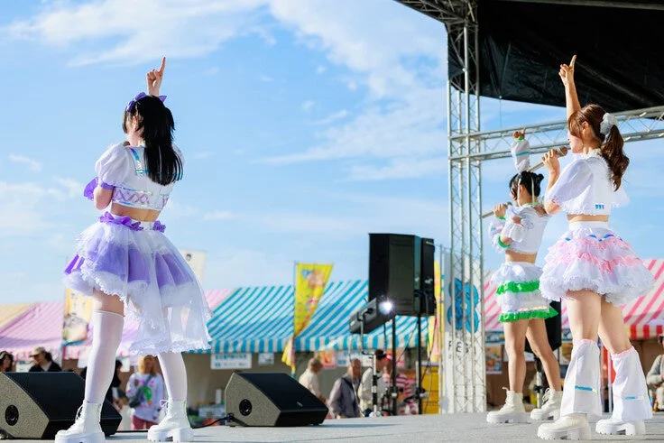 青空の下、屋外ステージでカラフルな衣装をまとった女性アイドルグループが歌い、パフォーマンスを披露している様子
