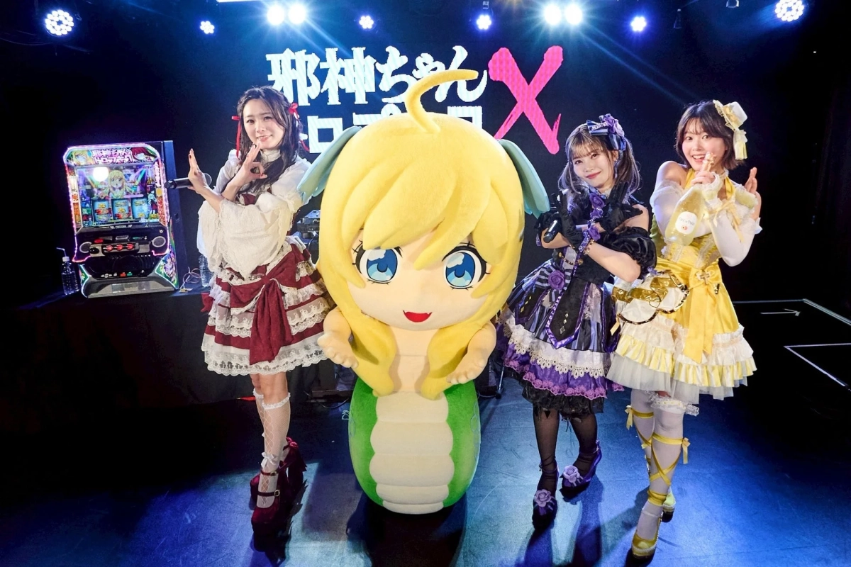 華やかな衣装をまとった3人の女性が、アニメ「邪神ちゃんドロップキック」のマスコットキャラクター「邪神ちゃん」の着ぐるみと共にステージ上でポーズをとっている。左にはパチンコ台が設置されており、イベントの様子を捉えたもの。