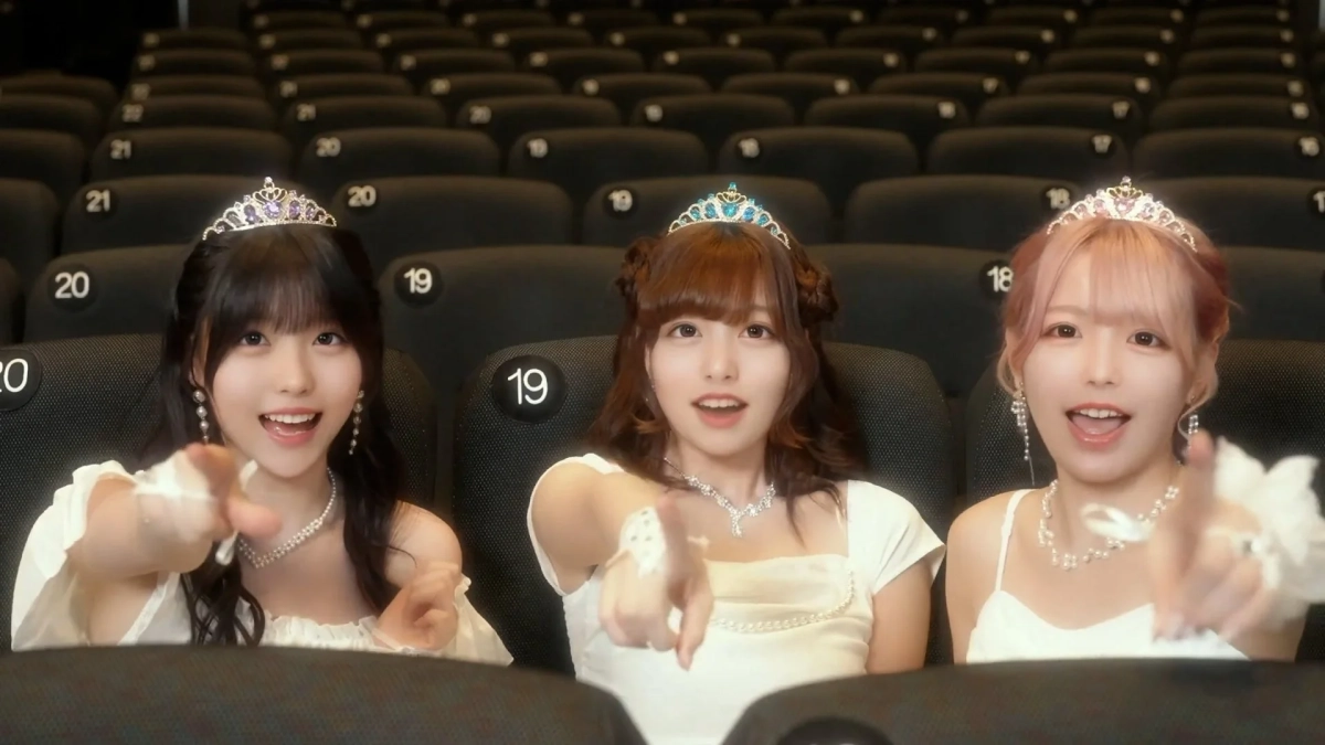 映画館のような観客席に座る3人の若い女性が写っています。全員ティアラと白いドレスを着用し、笑顔でカメラに向かって指をさしています。
